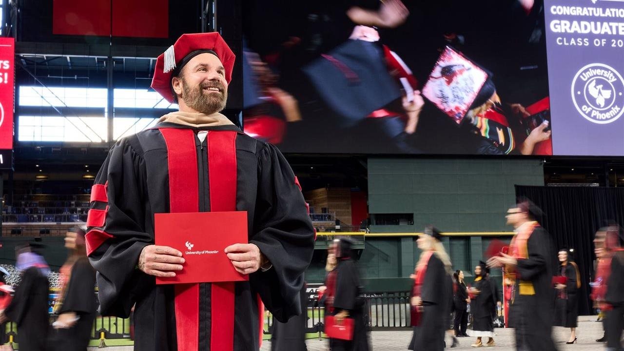 Man at a University of Phoenix graduation ceremony holds his diploma. University of Phoenix offers degree programs online.
