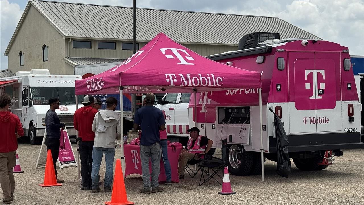 Un camión de servicios T-Mobile de color rosa brillante y personas de pie bajo una carpa instalada en la escena de un desastre.