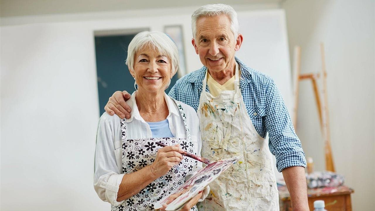 Pareja mayor, con paletas de pintura y delantales, disfruta de su capacidad de ver.  La American Society of Retina Specialists (ASRS) recomienda informarse más sobre la degeneración macular relacionada con la edad (DMAE) para proteger la visión.
