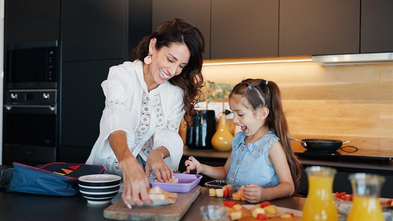 A woman and her daughter smile as they prepare box lunches in a kitchen.