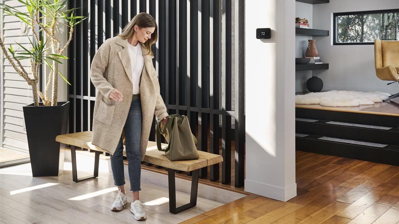Woman unbuttoning her coat and setting down her tote bag as she returns to her cozy, modern-style home, her Honeywell Home X8S thermostat set to 72 degrees.