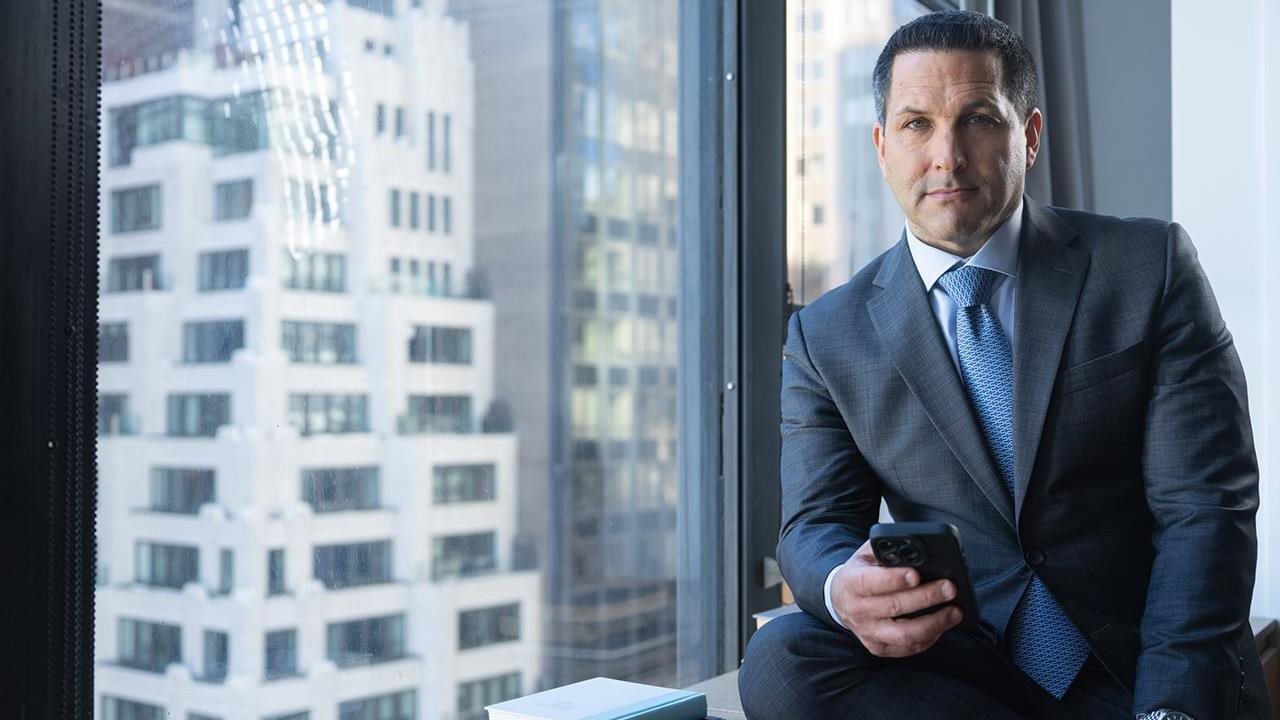 Adam wearing a blue suit and holding a cell phone while sitting on a window sill with large buildings in the background.