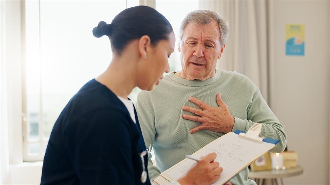 Nurse with a clip board talking with older male patient with hand on his chest.