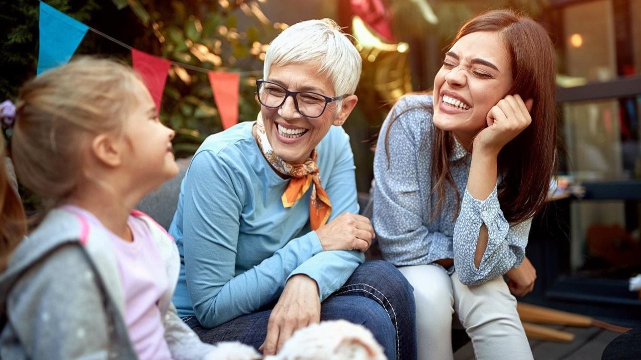 Grandmother, wearing glasses, laughing with daughter and granddaugter at a party.