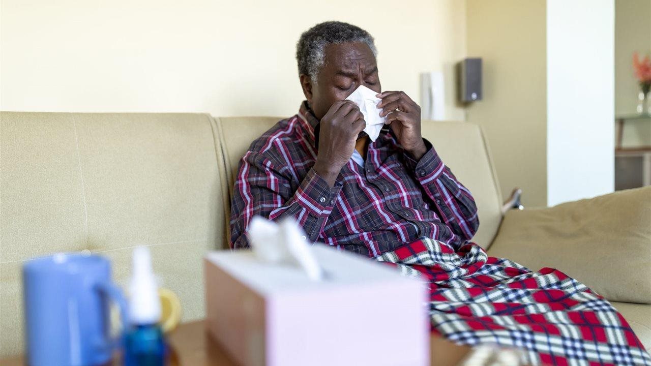 Sick senior man blowing his nose while sitting on the couch following the CDC advice by staying home and away from others until you're fever-free for at least 24 hours (without using fever-reducing medicines) and your other symptoms have improved.