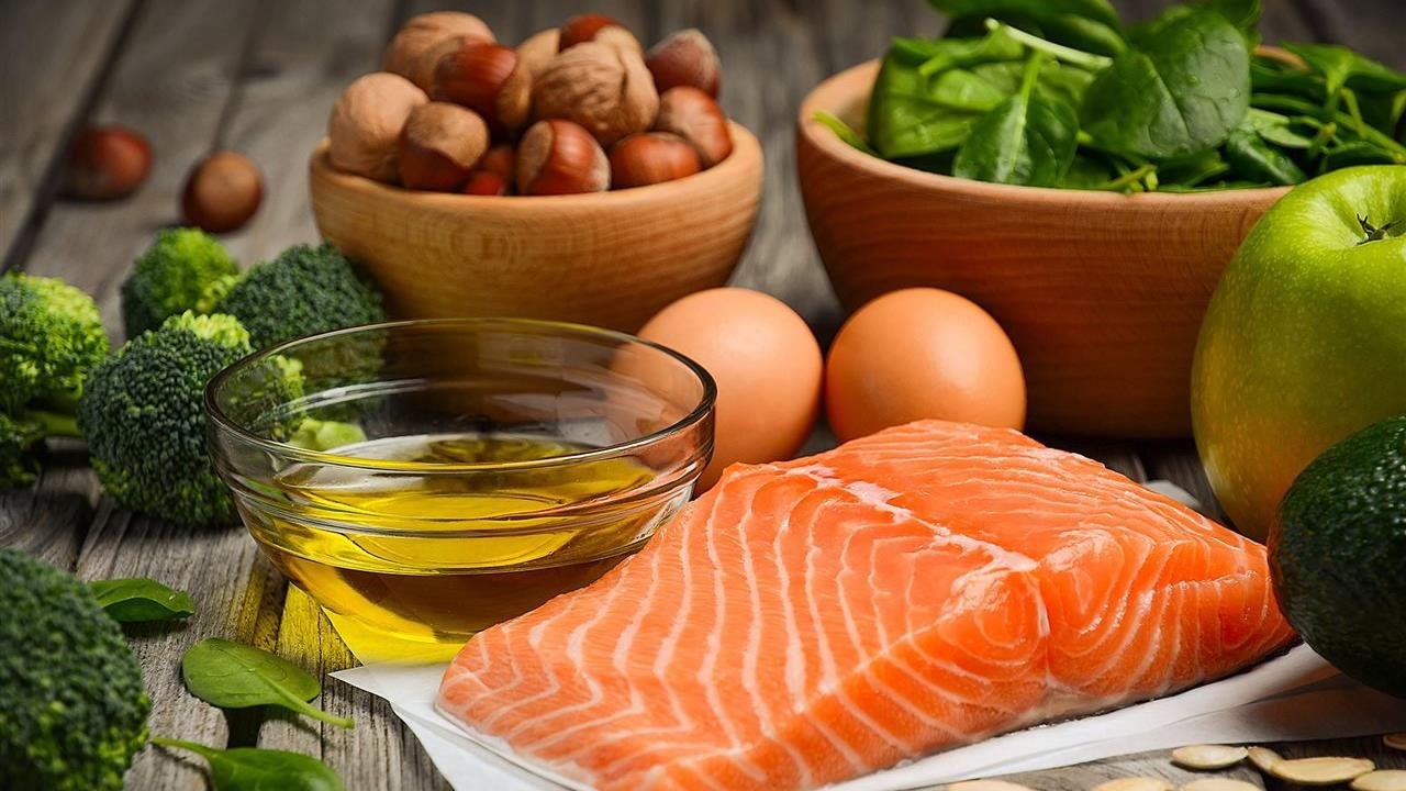 A salmon filet next to a small bowl of olive oil, two eggs, spinach, broccoli, apples and nuts.