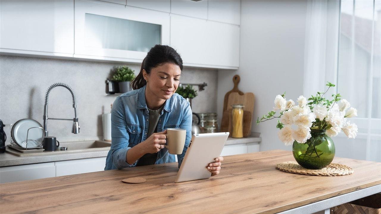 Woman drinking coffee while using tablet in kitchen. Navy Federal Credit Union has tips for staying safe online.