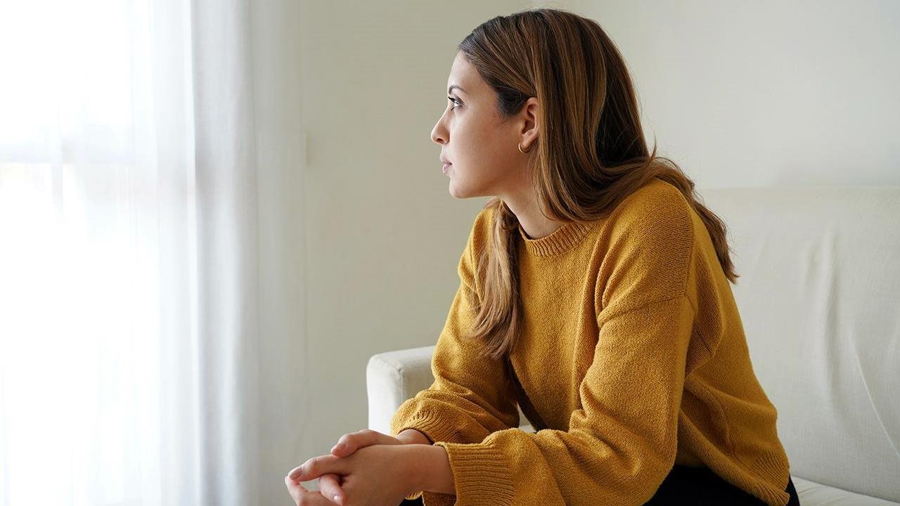 Una joven con un suéter amarillo está sentada en una habitación blanca, mirando por la ventana.