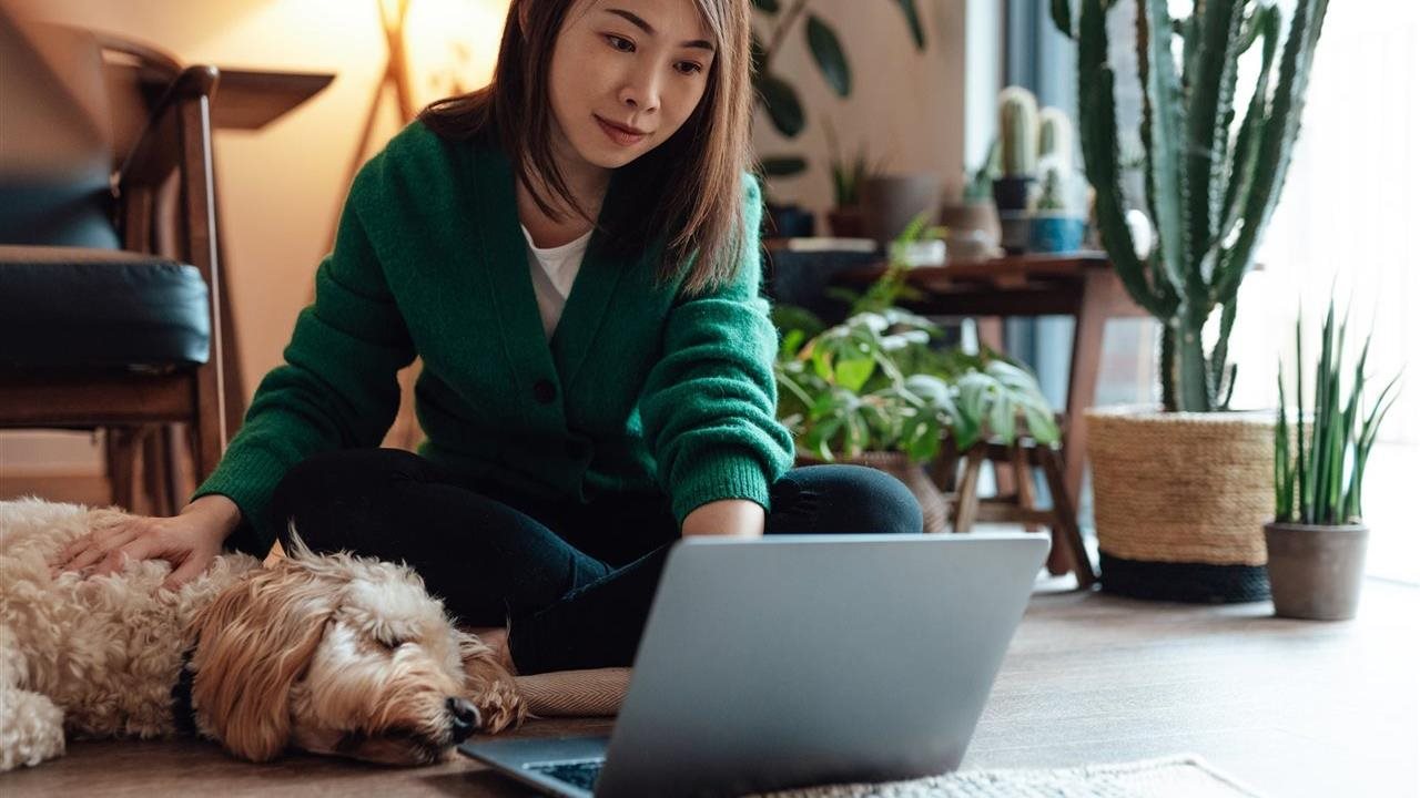 Young woman sitting on the floor of her apartment working on a laptop as her dog rests beside her.