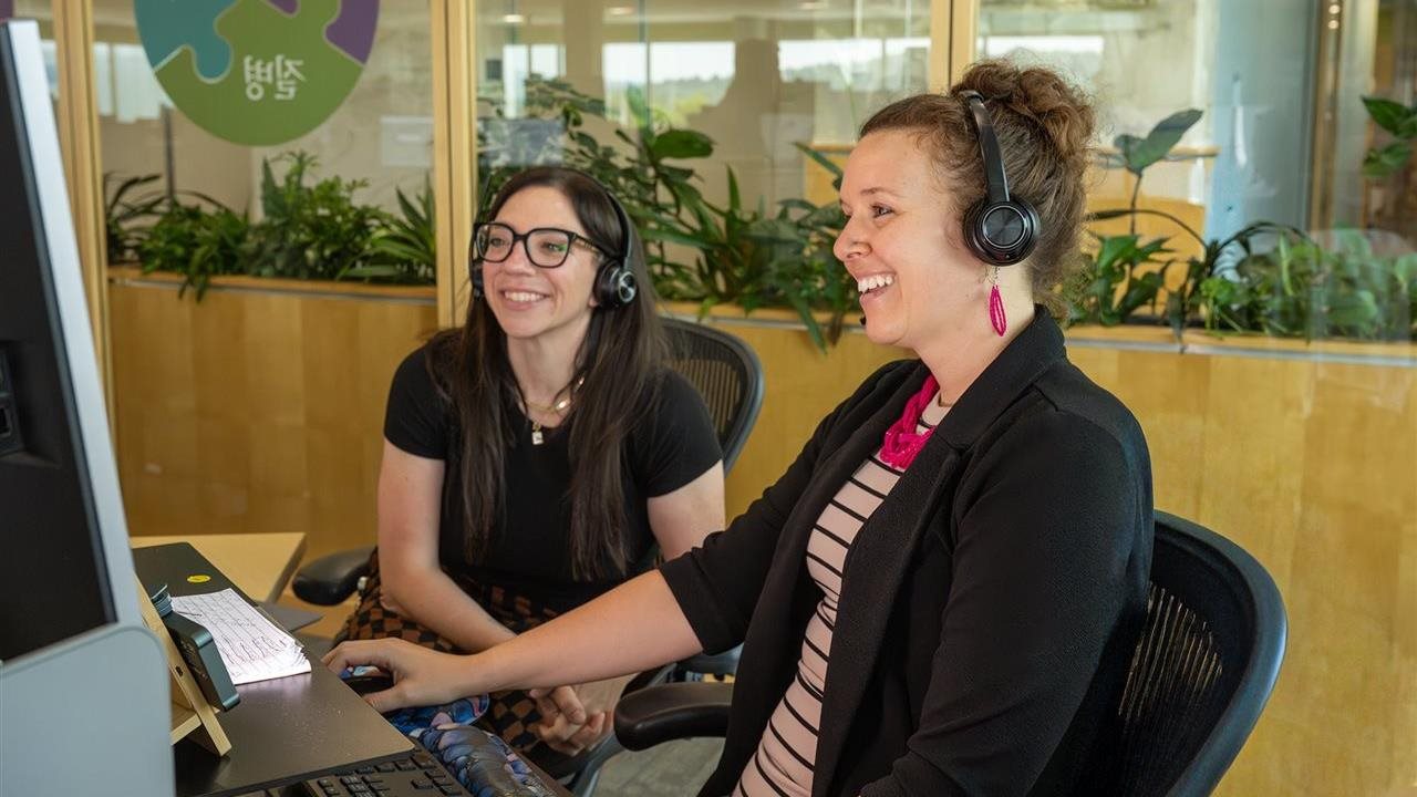 Dos mujeres sonrientes con audífonos sentadas frente a una computadora. El 2 de mayo es el Día Nacional del Seguro de Vida.