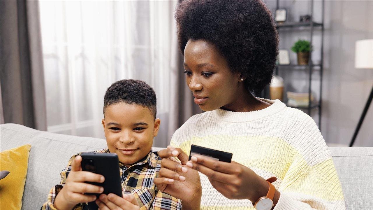 Mother with credit card and son with cell phone sitting on couch. ICCU youth accounts have free automatic parent transfers.