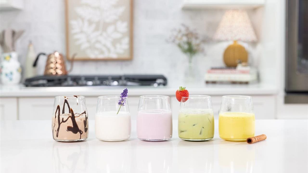 Glasses of flavored milk including honey lavender, strawberry matcha, golden, cherry vanilla moon and peanut butter banana mocha on a counter in the kitchen.
