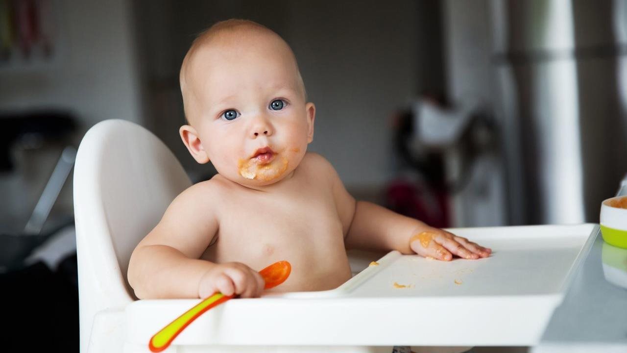Adorable baby with food on his face holding a spoon while sitting in a highchair.