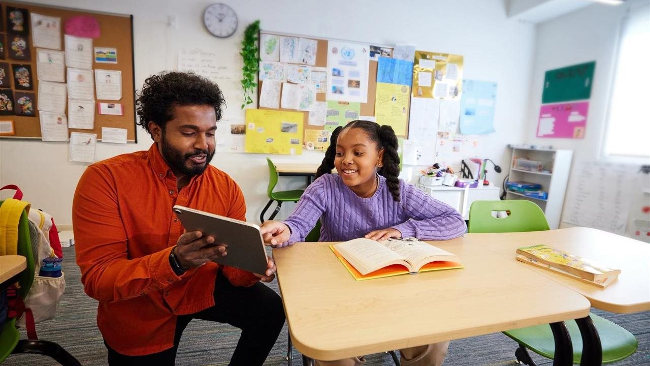 Female student in a classroom pointing to a tablet her instructor is holding.
