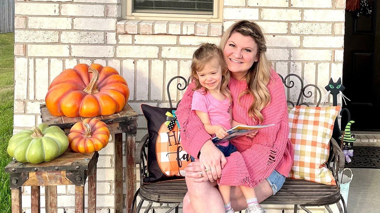 Megan sonriendo y su hija Kennedy, que murió de gripe, sentadas en muebles de patio al aire libre con decoraciones de otoño.