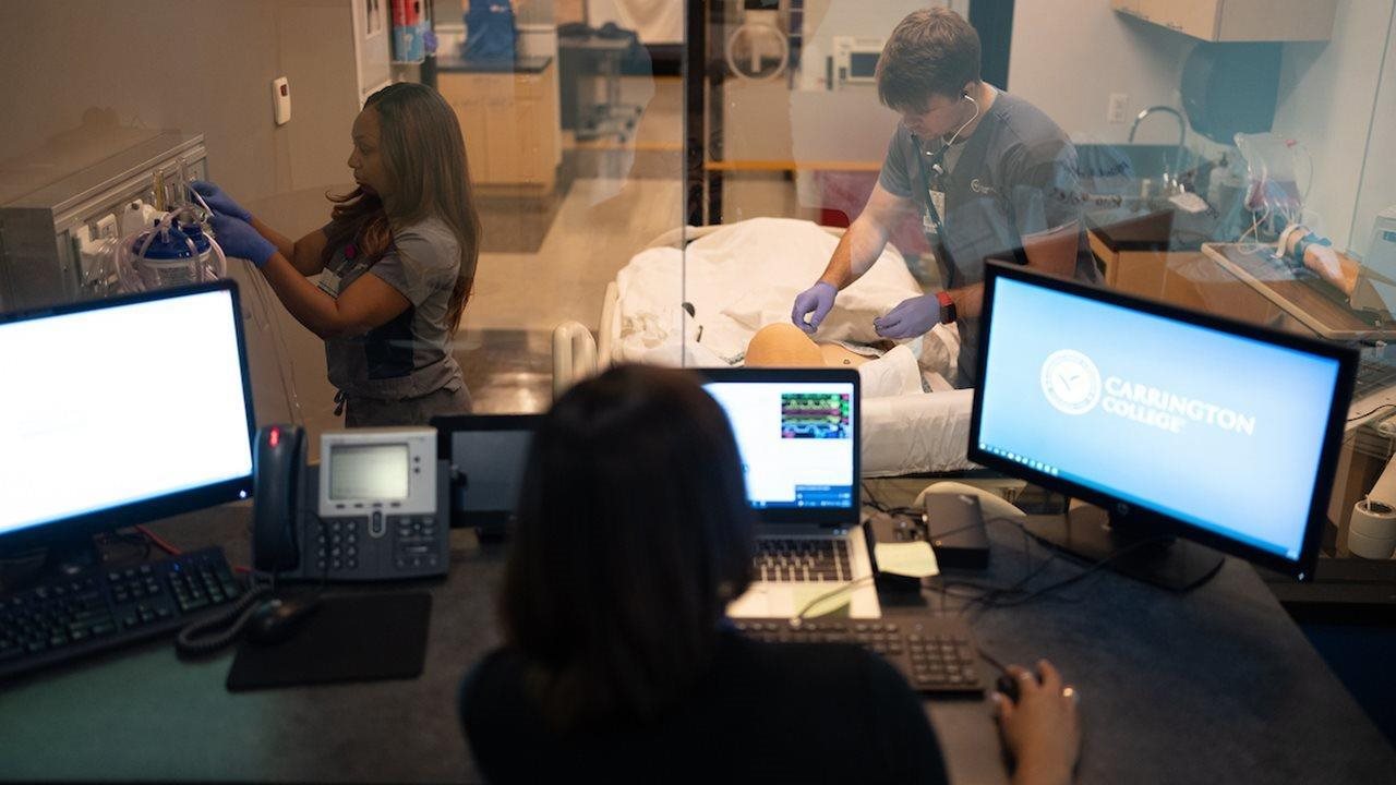Nursing students in an exam room praticing patient bedside care while another is studying online at Carrington College.