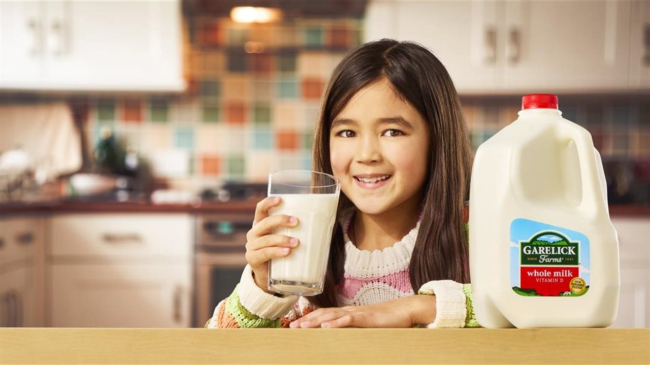 Young girl smiling with a glass of milk and a carton of Garelick Farms whole milk on the counter next to her.