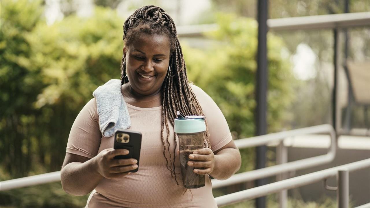 Woman using her phone and drinking water from a water bottle after a work-out in the gym.