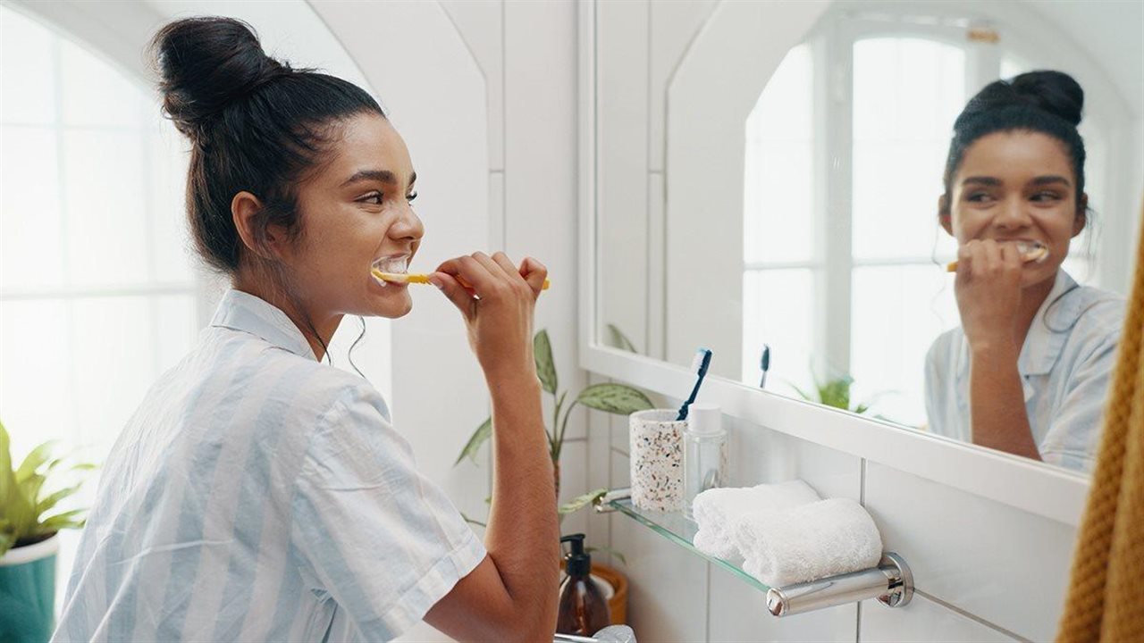 A young woman in pajamas brushes her teeth while looking in a mirror.