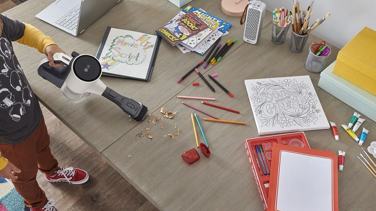 A boy using a hand vac to clean up a mess made by sharpening colored pencils on his desk at home.