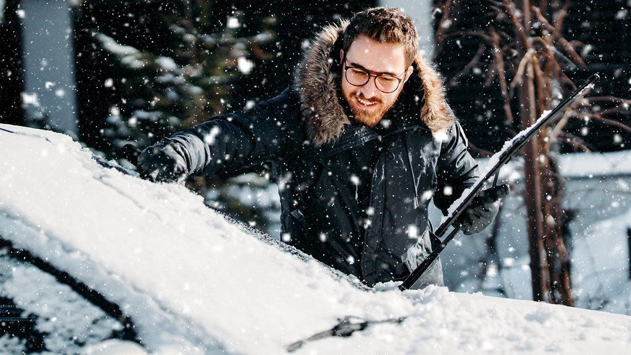 A man in a winter coat brushes snow off his windshield with his gloved hand.