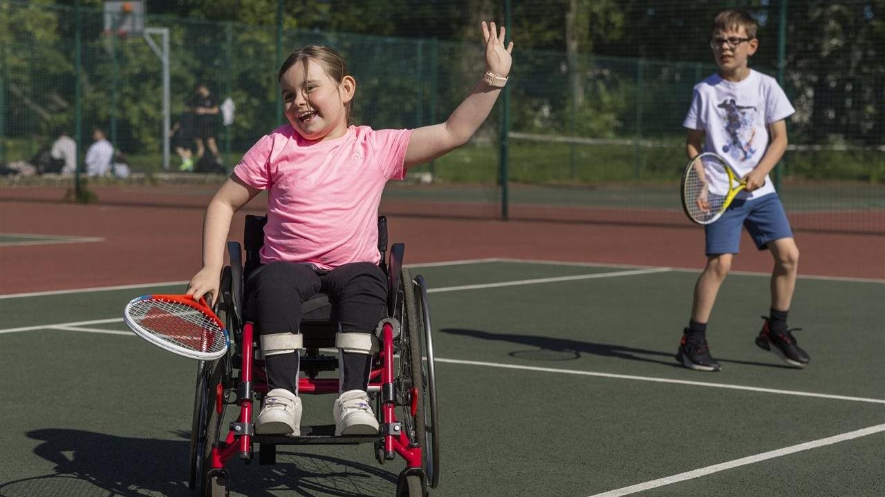 Little girl playing tennis from wheelchair with little boy in the background on the court.