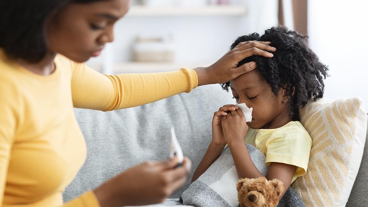 Mom taking her young daughter's temperature as she lays on the couch blowing her nose.