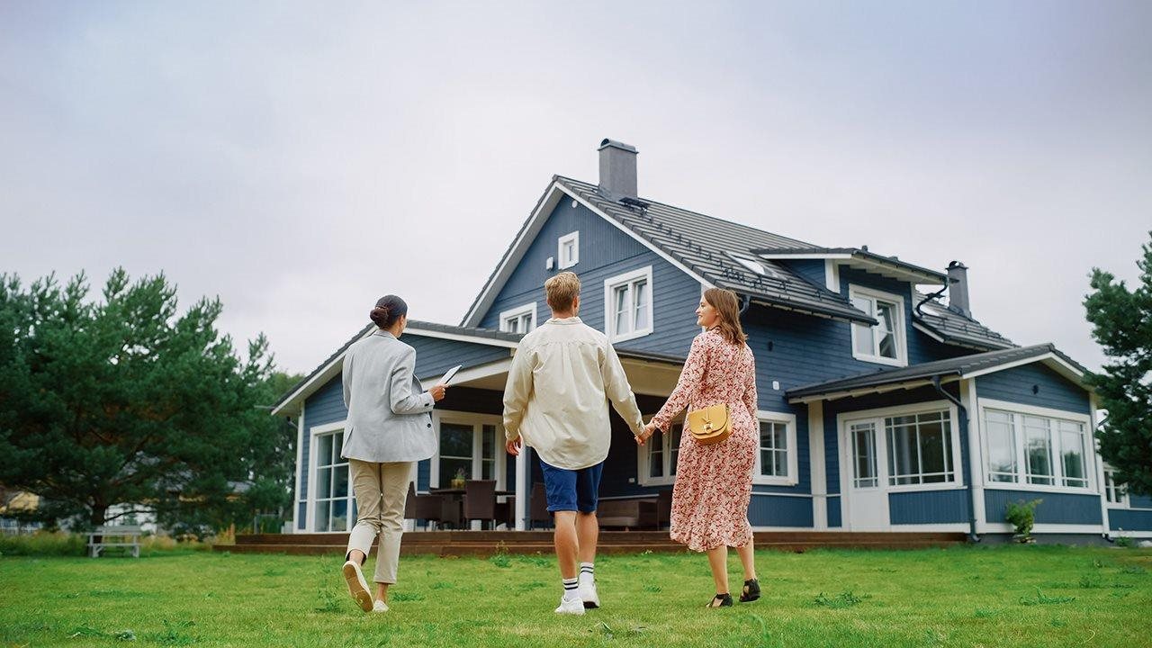 A Realtor walks a couple holding hands through the lawn toward a blue house.