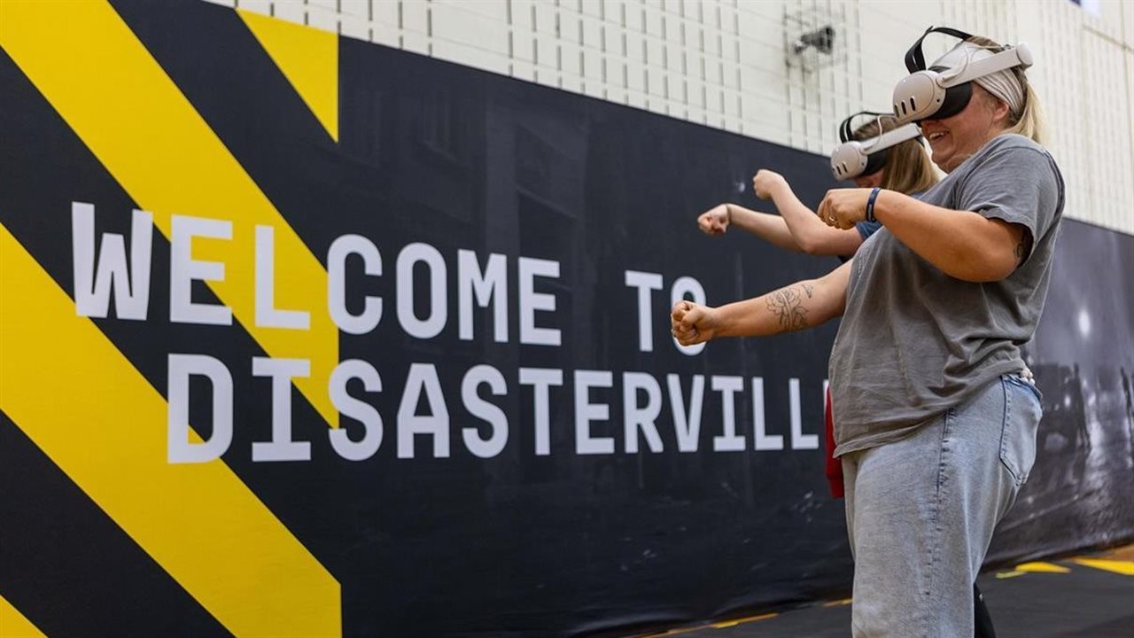 Two women wear VR headsets while standing in front of a wall reading "WELCOME TO DISASTERVILLE"