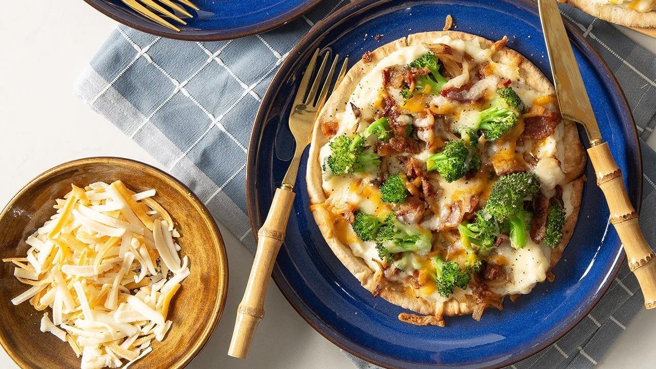 Mashed Potatoes pizza on the blue plate, next to a bowl of shredded cheese on a table.