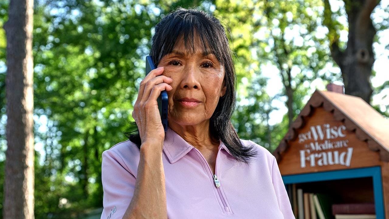 Senior woman on a cell phone outside next to a tiny library with a sign that says "Get Wise to Medicare Fraud".