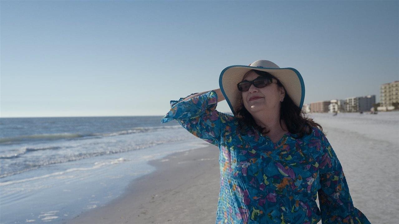 Smiling Kathy, living with urothelial cancer, in a floppy hat and sunglasses walking along the beach. Once Kathy's diagnosis was confirmed as LG-UTUC, her doctor recommended JELMYTO®