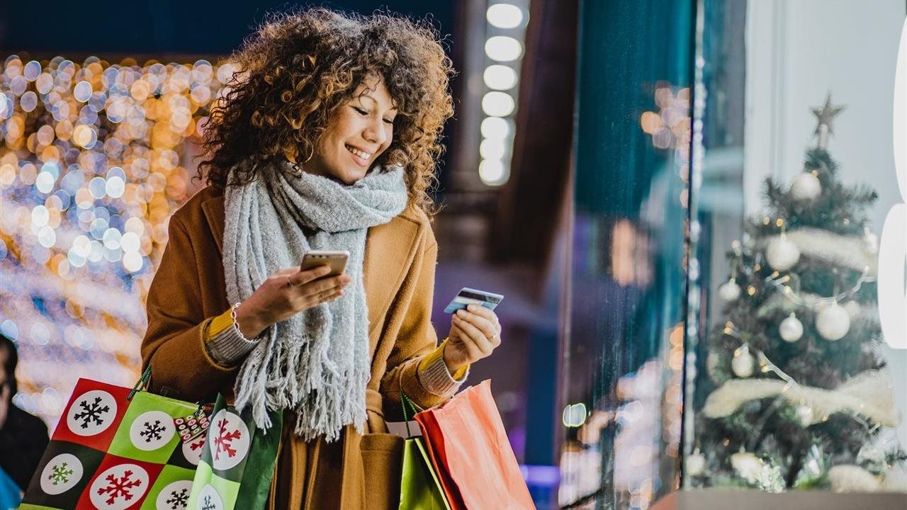 Mujer sonriente con abrigo de invierno y bufanda sosteniendo bolsas de compras y usando su teléfono celular mientras compra regalos afuera de la ventana de la tienda.