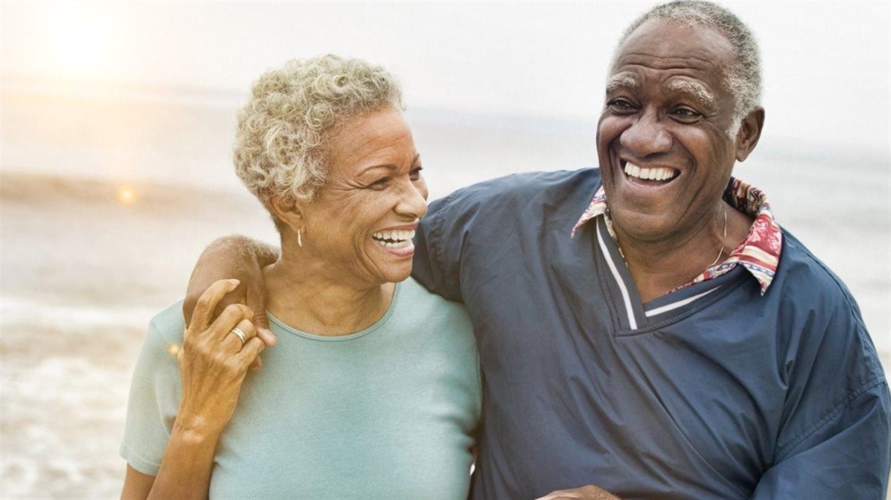 A senior Black couple smiles as they embrace walking along a beach at sunset.
