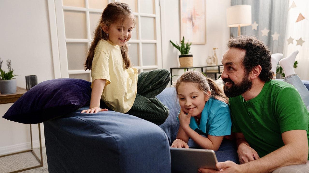 A dad and his two daughters lay on a couch, looking at their tablet