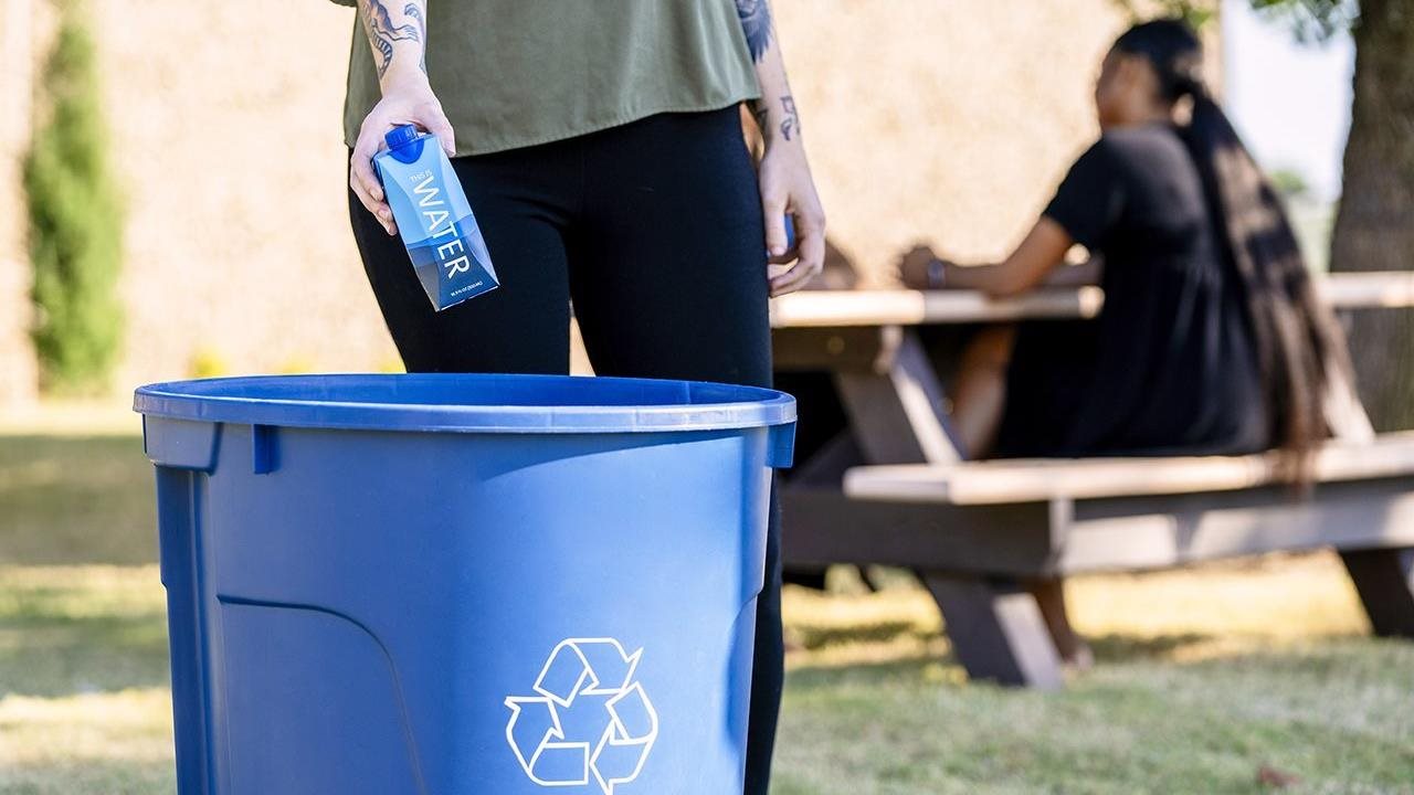 Woman tossing paper water carton into recycling bin.