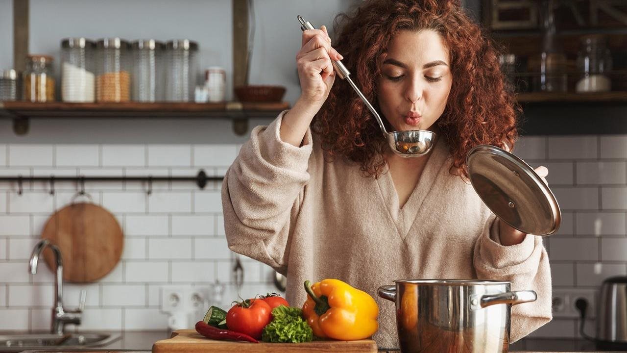 Woman blowing on soup in a ladle over a pot before trying a new dish she is preparing in the kitchen