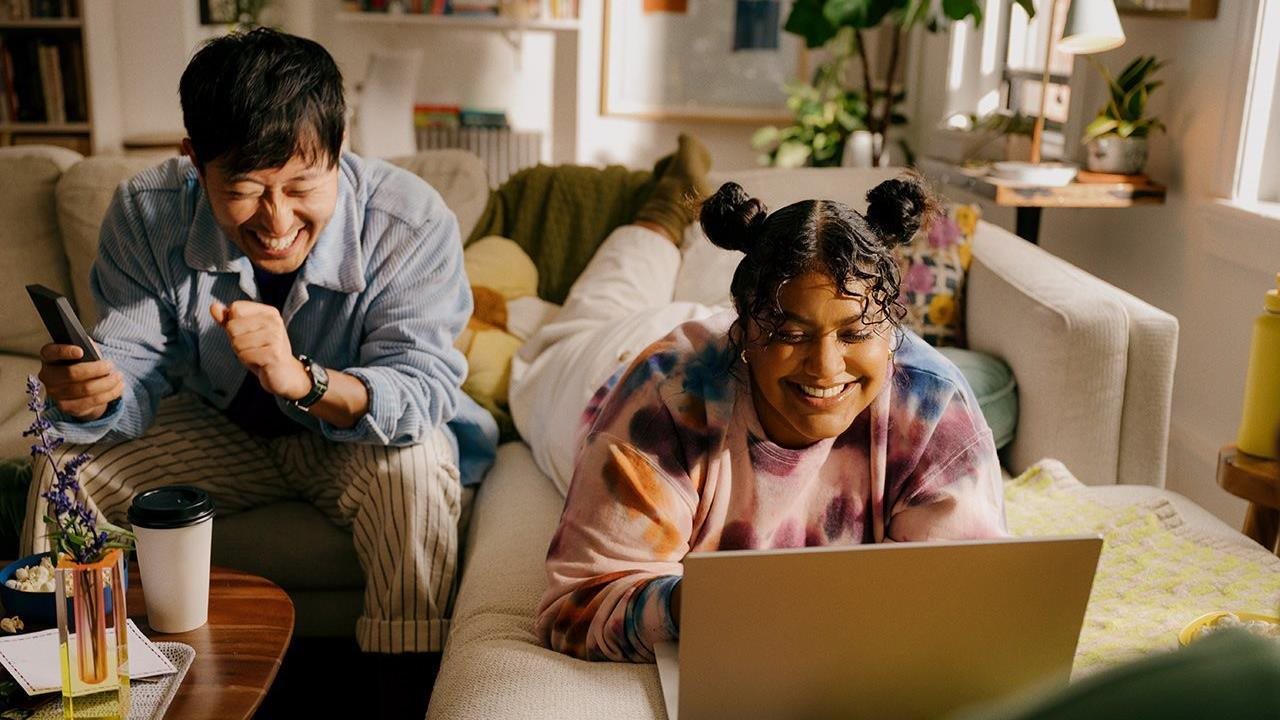 A man and woman laugh while relaxing on their couch, watching a laptop computer together.