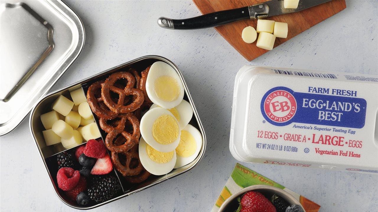 A snack tin with sliced boiled eggs, pretzels, cheese and fruit next to a carton of Eggland's Best eggs.