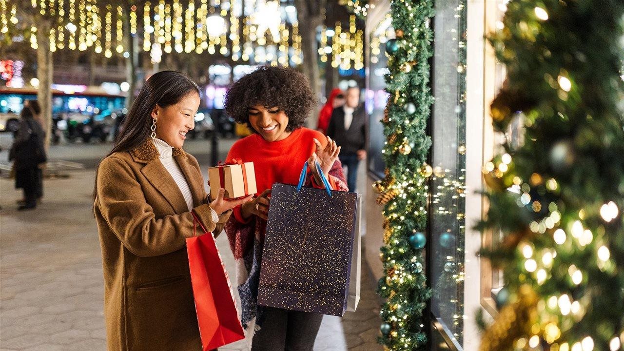 Two women holiday shopping at night on a main street with many shops and holiday lights.