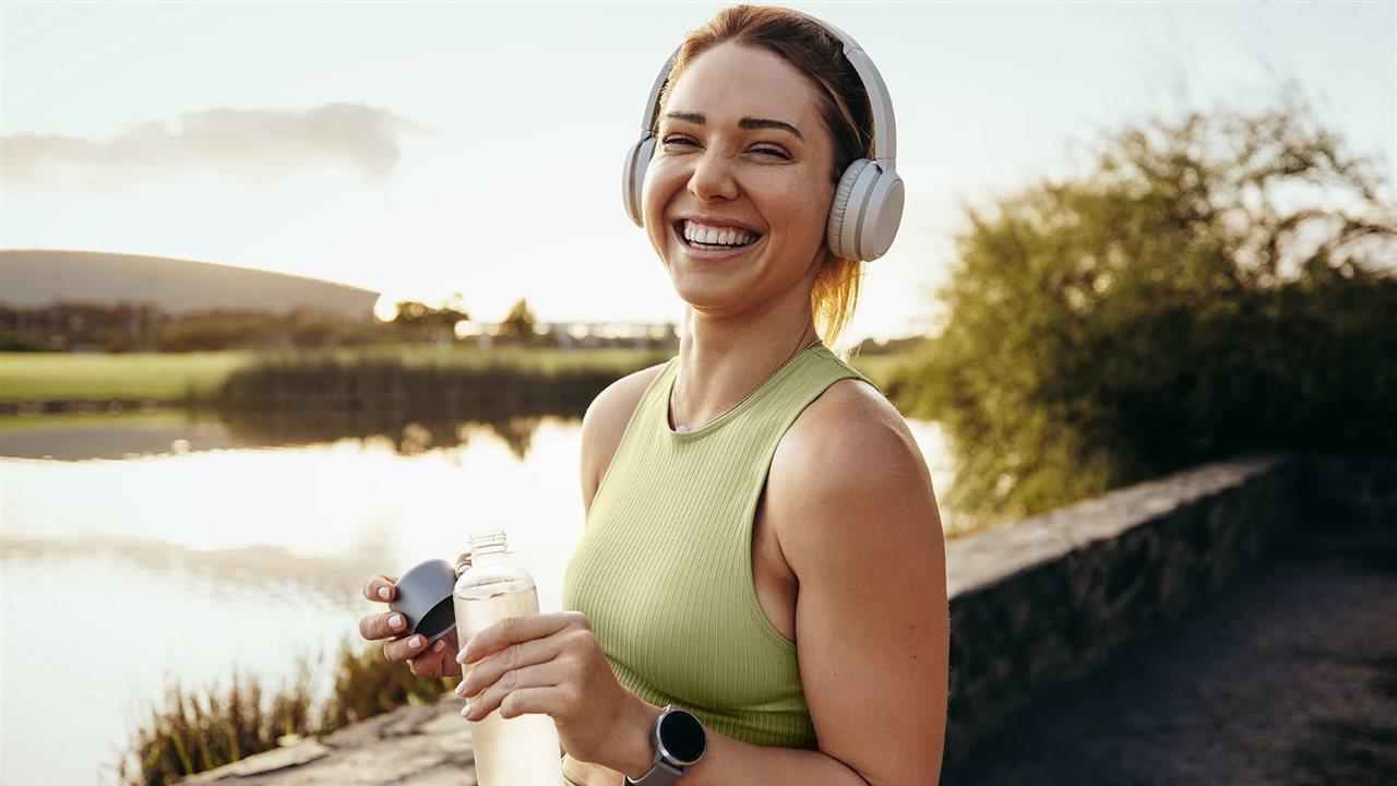 Smiling woman wearing headphones and drinking water while enjoying a view of the lake.