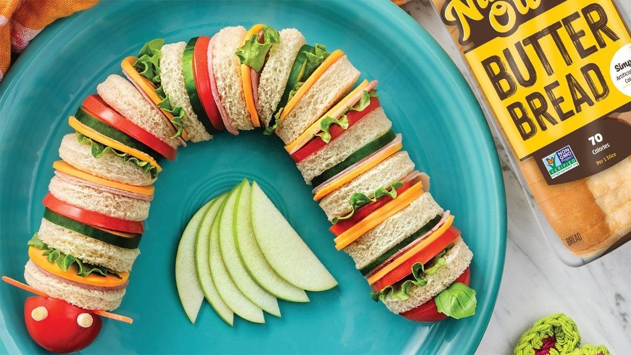 Caterpillar shape sandwich with cucumbers, cheese, meat and lettuce plated next to a loaf of Nature's Own Butter Bread.