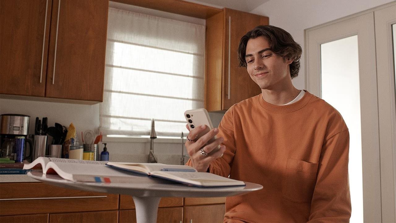 A young man smiles as he looks at his phone while sitting at a kitchen table.