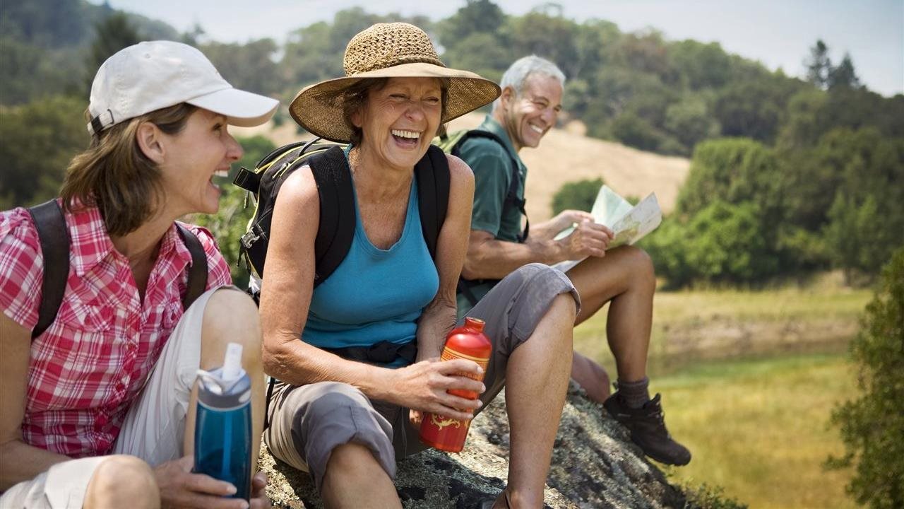 Group of older friends pausing their hike to drink water while sitting on a large rock.