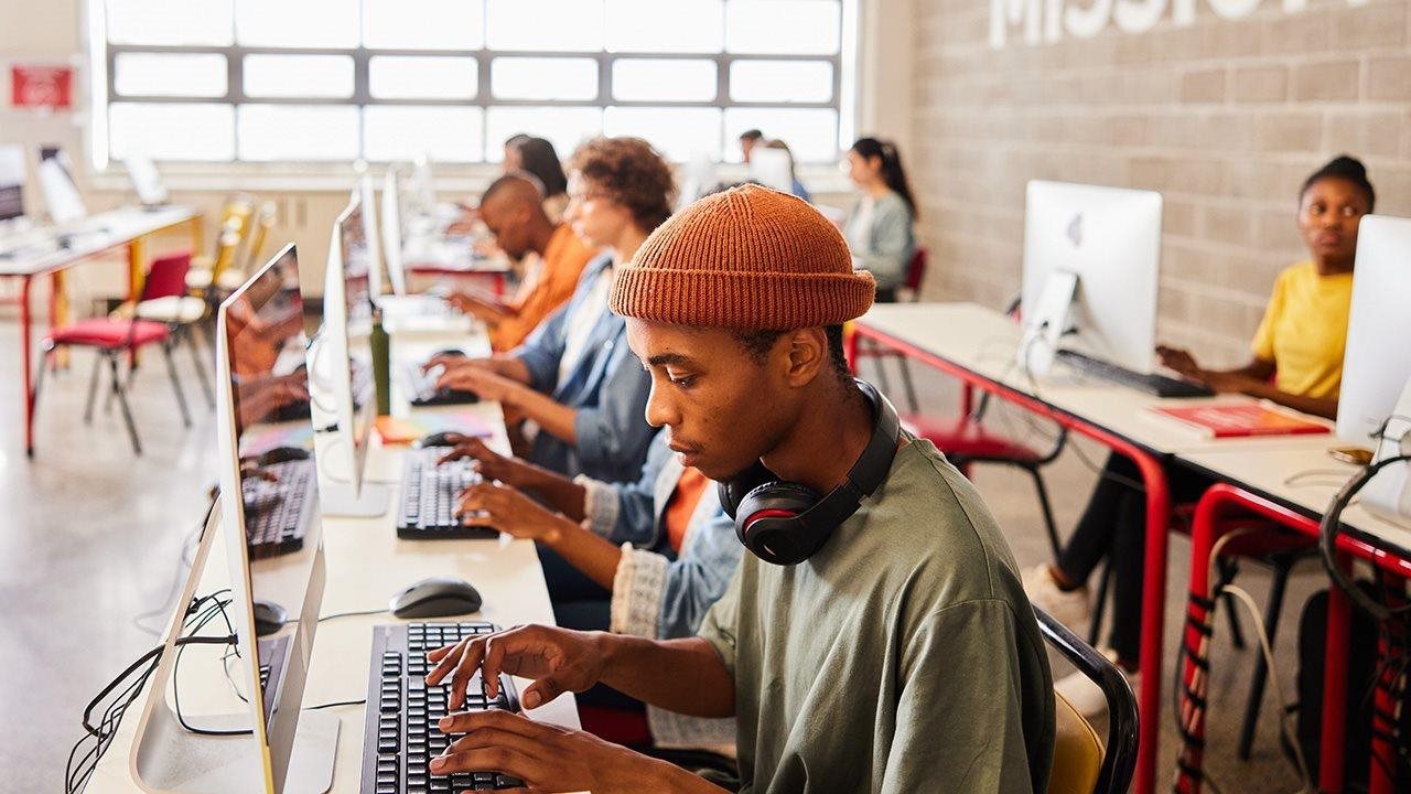 A young Black man wearing a knit orange cap works at a desktop in a computer lab.