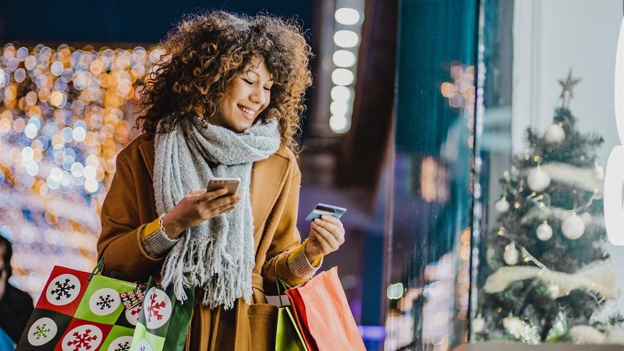 Smiling woman wearing winter coat and scarf holding shopping bags and using her cell phone as she shops for gifts while outside store front window.