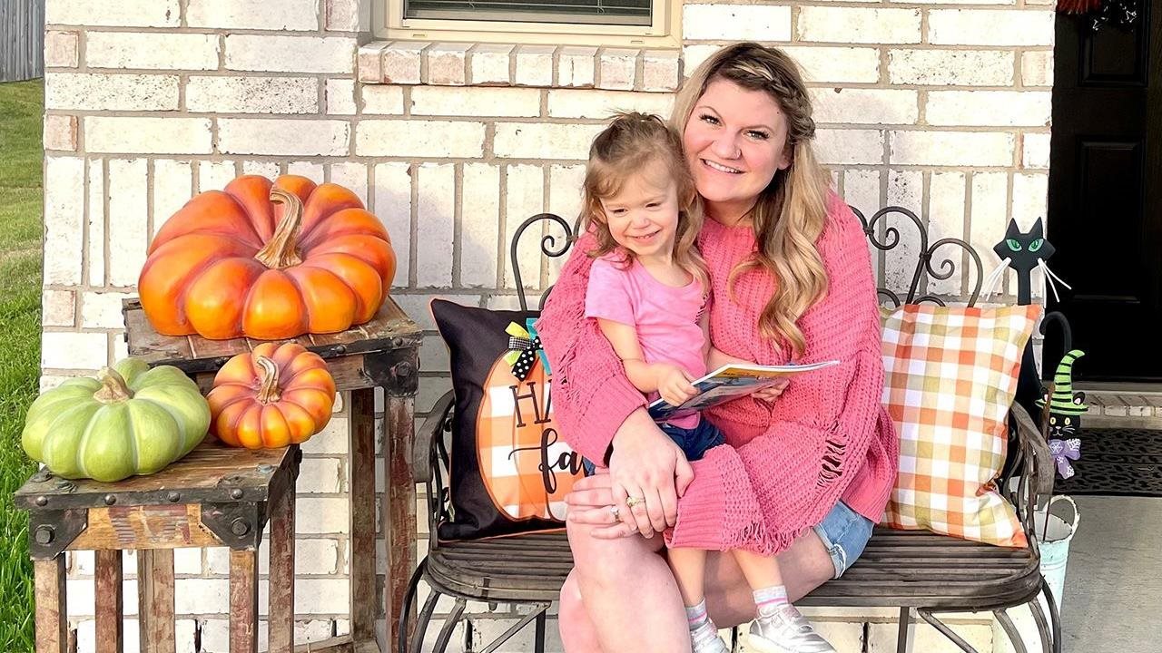 Smiling Megan, and her daughter Kennedy who died from the flu, sitting on patio furniture outside with Fall decorations.