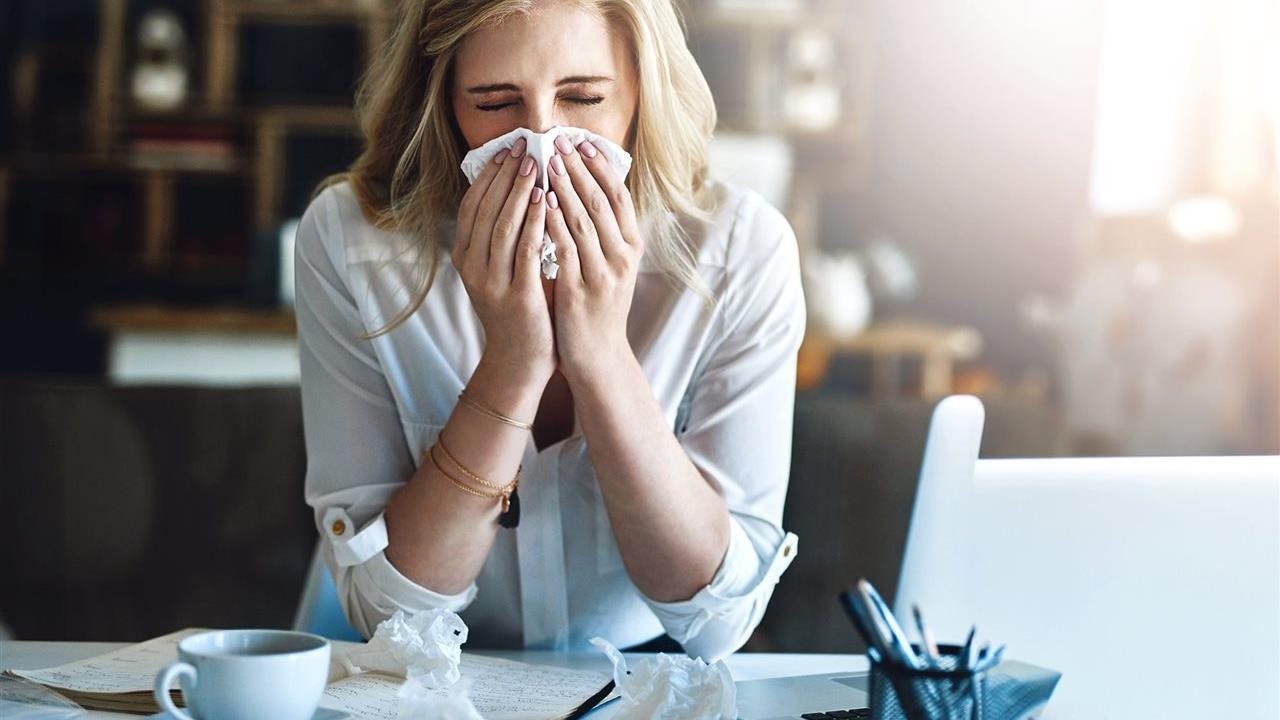 Woman at her desk sneezing as she is attempting to get back to work after a cold.