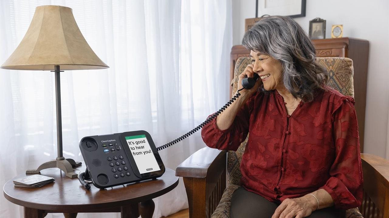 Senior woman sitting in a chair in her living room talking with a friend using a ClearCaptions telephone.