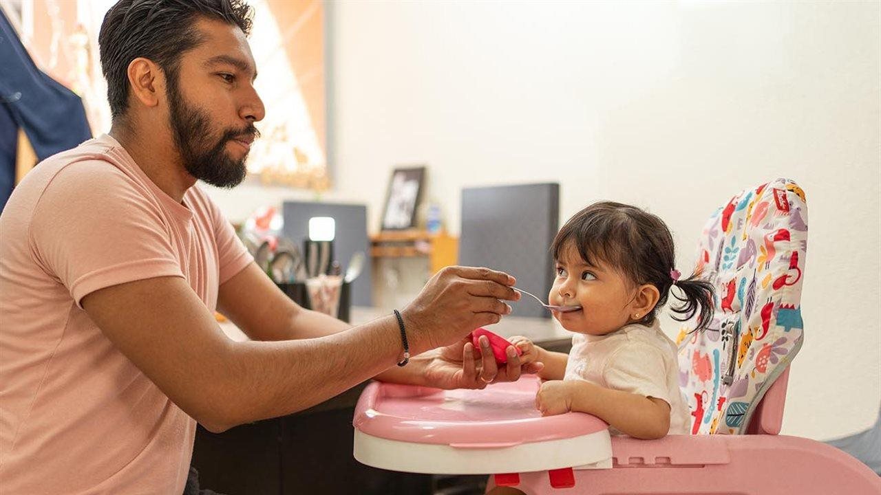 Papá alimentando a su hija pequeña de un tazón pequeño mientras ella está sentada en una silla alta.
